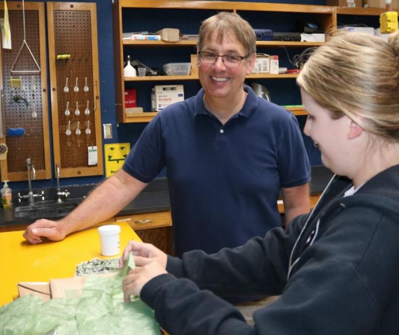 WIST executive director Paul Fowler works in the compostability lab at WIST. He is smiling at the camera while a woman employee from WIST shows his plastic samples.