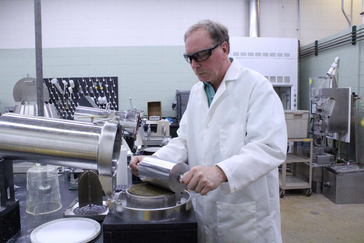 A man in a lab coat is holding a roller while making paper hand sheets for paper testing.
