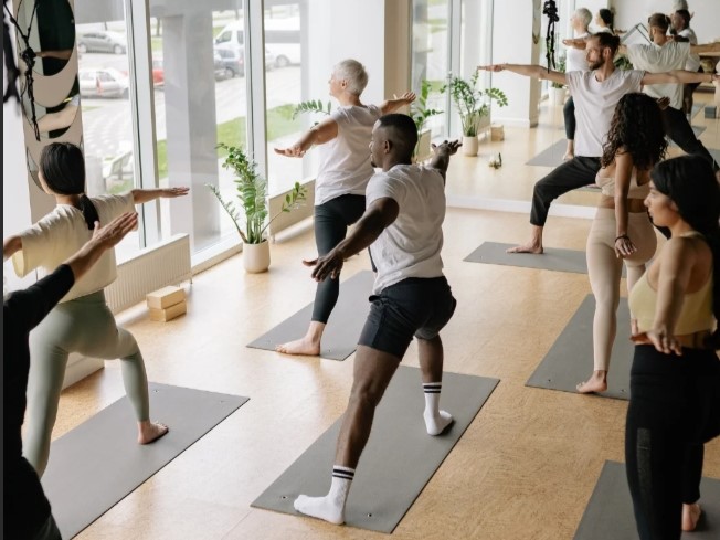A group of people practice yoga poses on mats, facing large windows with bright sunlight streaming into the room.