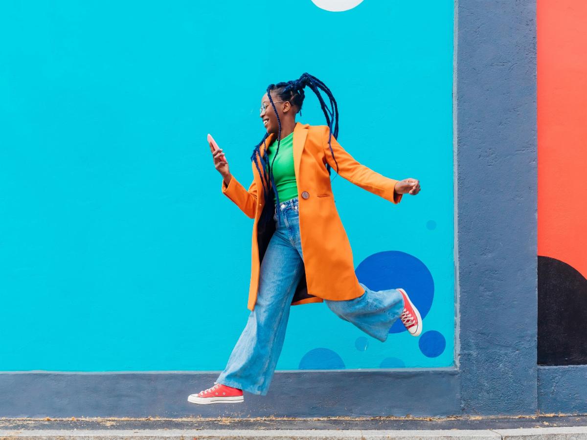 Women in brightly colored clothing jump in the air while smiling and holding a cellphone against a colorful background.