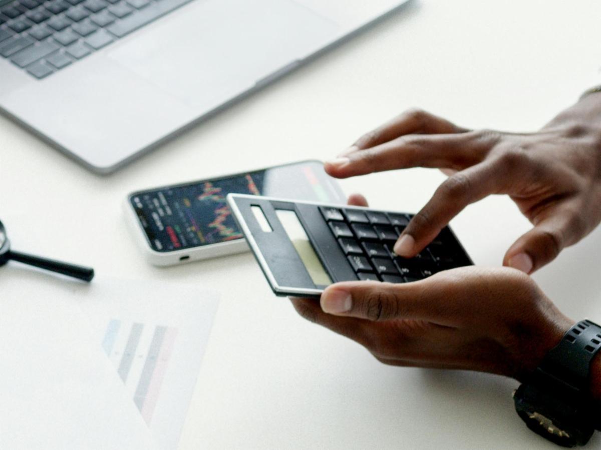 Hands use a calculator with a smartphone and laptop visible in the background.