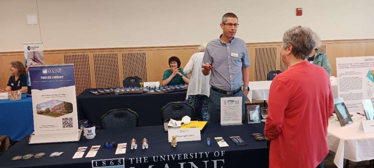Photo of Fogler table at Retiree Homecoming. Matthew Revitt speaks to an attendee.