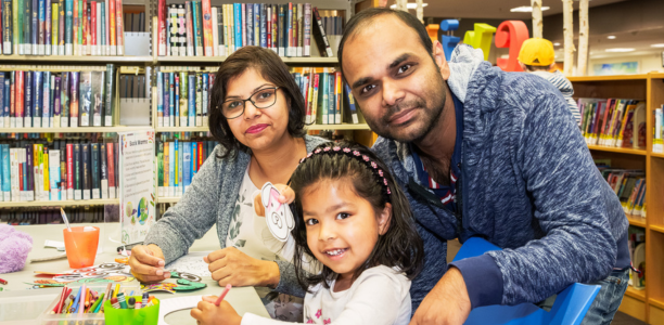 : A family smiles while doing a children’s craft activity together at a library table, with bookshelves visible in the background.