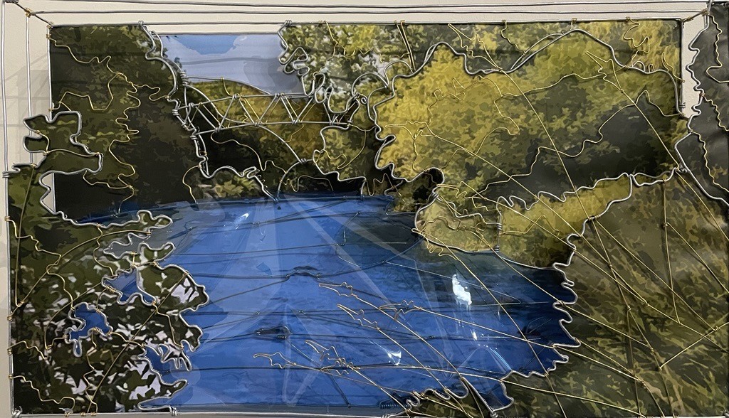 A mixed‑media artwork depicts a tranquil blue river winding through dense green foliage on both sides, overlaid with raised, contour-like lines and intersecting marks that suggest a mapped landscape, all framed behind glass.