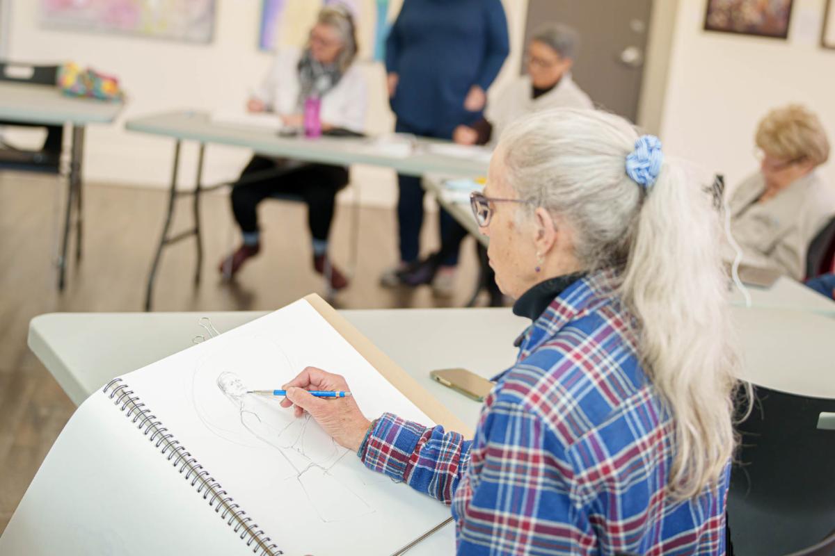 A person seated at a table sketches in a large drawing pad while others sit and observe in the background of an art classroom.