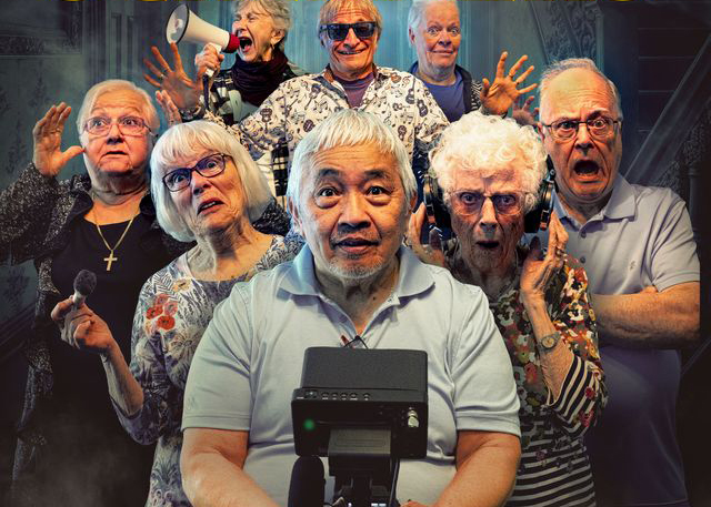 “A group of people stands closely together in a dramatic, dimly lit setting that resembles a staged scene or poster. Some individuals hold props such as a megaphone, a microphone, and a camera monitor. The group is arranged in multiple rows, with one person seated in front operating a piece of video equipment. The background includes dark, atmospheric architectural details, adding to a theatrical or cinematic mood.