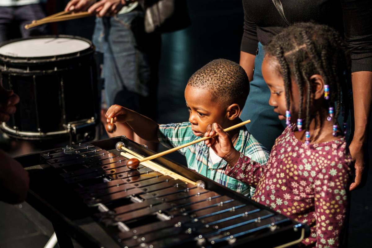 Children playing a xylophone with mallets while standing close together on a stage with a drum visible in the background and an adult nearby.