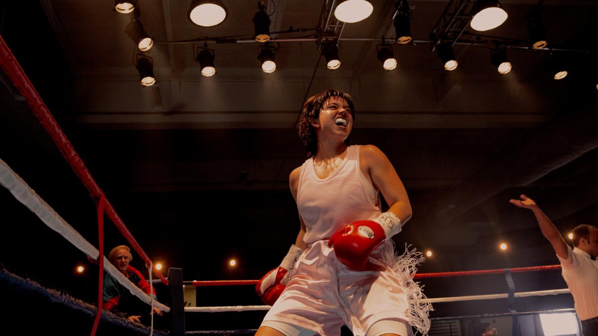 A boxer wearing a white athletic outfit and red gloves stands inside a boxing ring under bright overhead lights. The boxer is positioned near the ropes, leaning slightly forward with hands raised. Another person is visible at the edge of the ring with an arm extended upward, and additional figures appear in the background of the dimly lit arena.