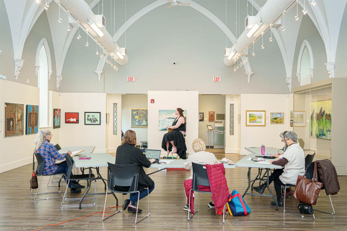 Interior view of an art gallery with a high, arched ceiling, white walls, and track lighting. Six people are seated at rectangular tables arranged in a semicircle on a wooden floor, drawing or taking notes on paper and laptops. At the center, a nude model sits elevated on a red platform, posed against a temporary white wall displaying framed artwork. Framed paintings and drawings hang along the side walls, and exit signs are visible above doorways at the back of the gallery.