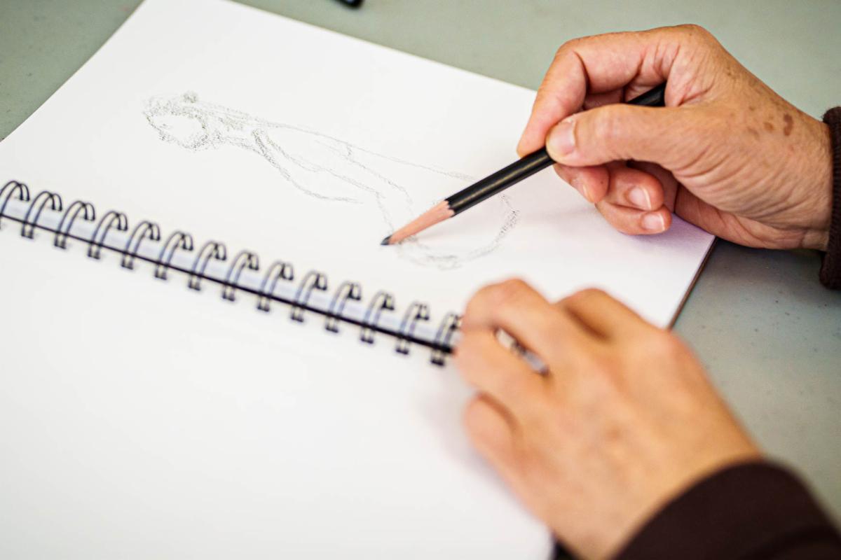 Hands holding a pencil sketching a figure on a spiral bound sketchbook.