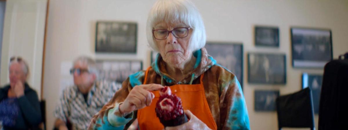 A person with short light-colored hair wears an orange apron and holds a partially carved red vegetable or fruit, using a small carving tool. They are in a room with framed photographs on the wall behind them, and other people are seated in the background.