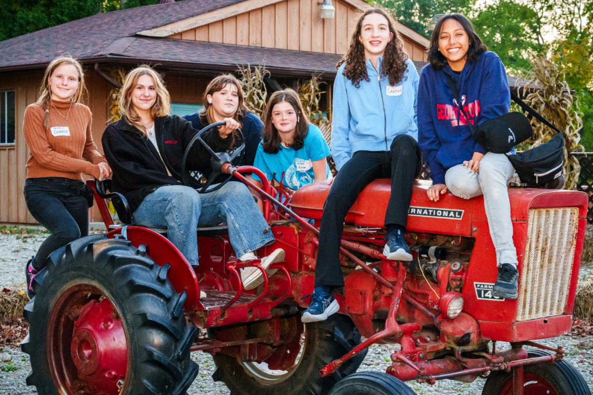 Six people sit and lean on a red tractor parked outdoors in front of a wooden building. Trees and greenery surround the scene, and the group wears casual clothing, with backpacks and the tractor’s large front tire visible in the foreground.