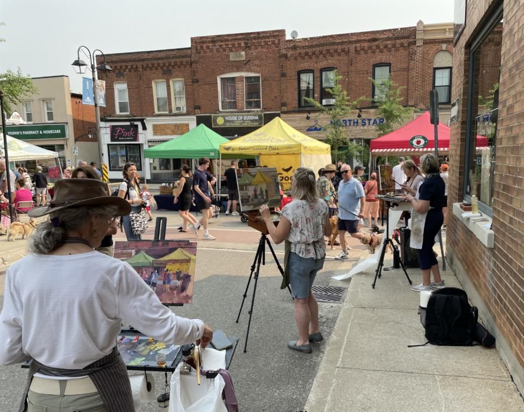 Several artists paint outdoors along a closed main street during a plein air art event, with easels and tripods set up on the sidewalk as pedestrians pass by vendor tents and brick storefronts in the background.