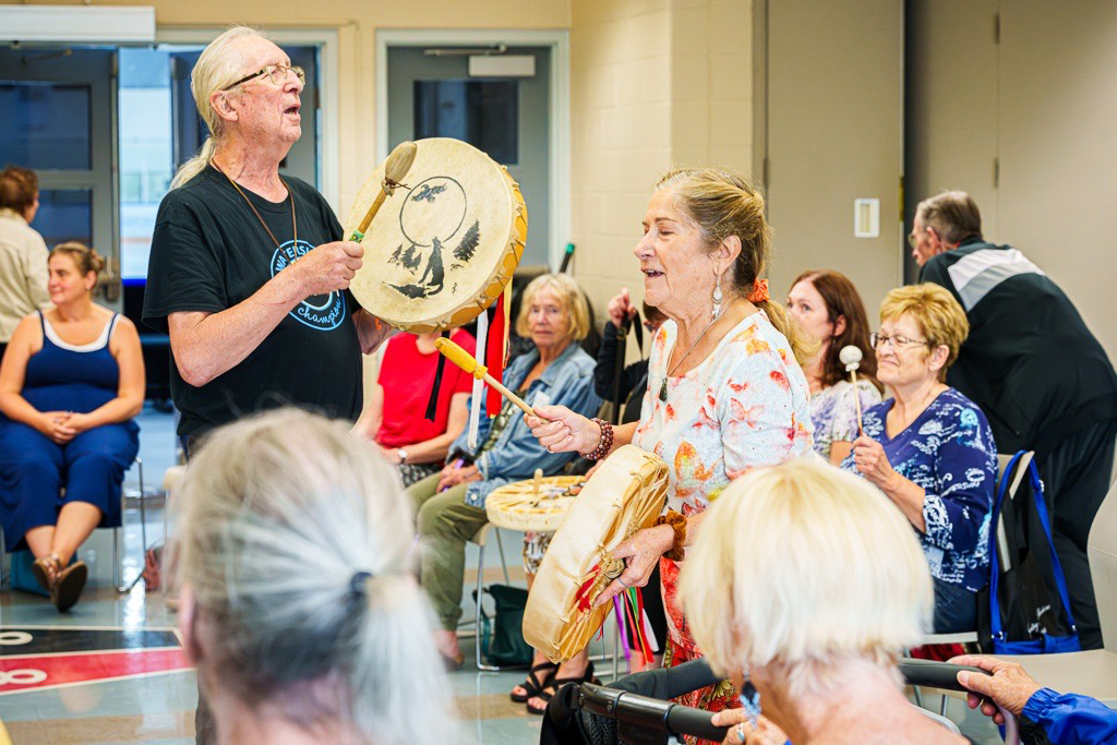 Indoor gathering with several people seated and standing in a room. Two Indigenous individuals in the center hold large hand drums and drumsticks. Other participants sit in chairs arranged in a semicircle. Some people hold smaller percussion instruments. The setting is a community or cultural event with bright lighting and neutral walls. The floor has a patterned design with red and gray tones.
