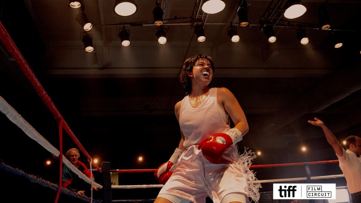 A boxer wearing a white athletic outfit and red gloves stands inside a boxing ring under bright overhead lights. The boxer is positioned near the ropes, leaning slightly forward with hands raised. Another person is visible at the edge of the ring with an arm extended upward, and additional figures appear in the background of the dimly lit arena. The TIFF Film Circuit logo is placed on top the image in the bottom right corner.