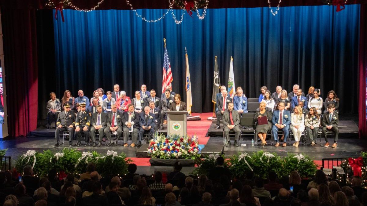 Wide shot of an auditorium stage with state and local elected officials seated as a speaker stands at the podium.