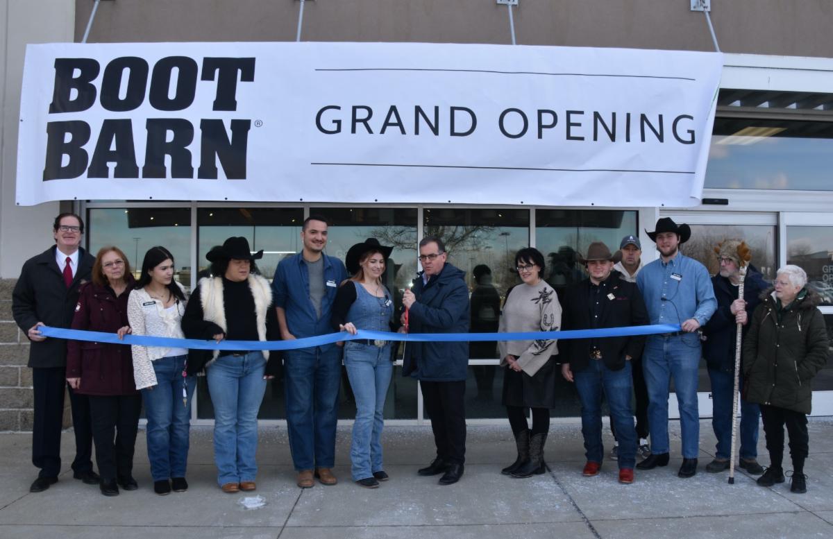 A ribbon-cutting ceremony takes place outside the Boot Barn store, with Mayor Robert Van Campen and community members standing in a line holding a blue ribbon beneath a large banner that reads “Boot Barn Grand Opening.” Several people wear Western-style hats and denim, and the storefront entrance is visible behind them.