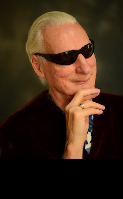 The generational leader, Mr. Larry with slicked-back white hair, wears dark sunglasses and rests his chin on his hand smiling thoughtfully against a soft, blurred background.