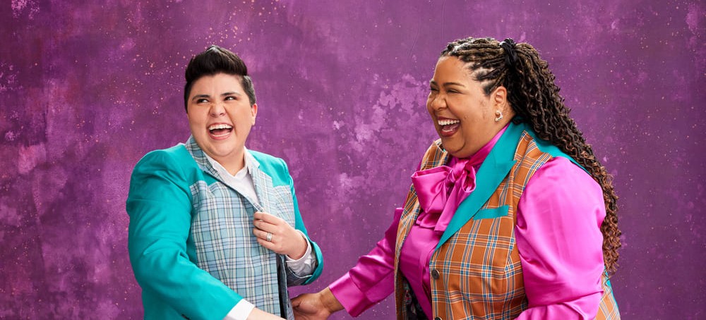 Two women smiling and laughing wearing colorful suits in front of a purple background