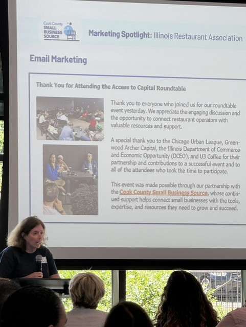 Presenter speaks into a microphone before an audience as a projected email marketing slide thanks attendees of the Access to Capital Roundtable and highlights event partners.
