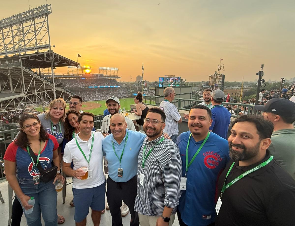 Group of attendees smiling for a photo on the Wrigley Rooftops overlooking Wrigley Field during a Chicago Cubs game at sunset. The baseball field, packed stadium, and Chicago skyline create the backdrop for the networking event.