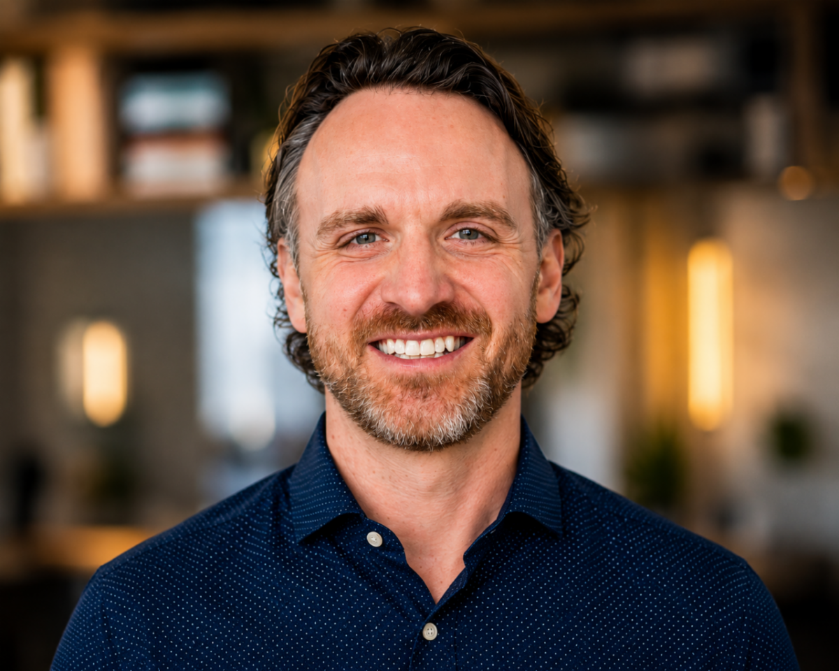 Smiling man with short dark hair and a beard, wearing a navy blue button-down shirt against a softly blurred indoor background.