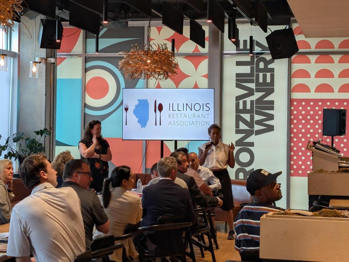 A speaker addresses seated attendees at a restaurant event as another woman stands nearby. A screen displays the Illinois Restaurant Association logo against colorful Bronzeville Winery wall graphics.