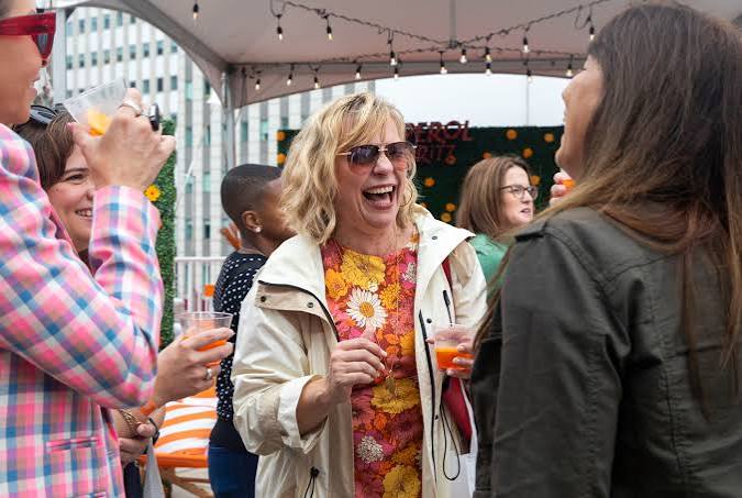 A woman in sunglasses and a floral dress laughs while holding a red drink and talking with a group of adults at an outdoor gathering under string lights.