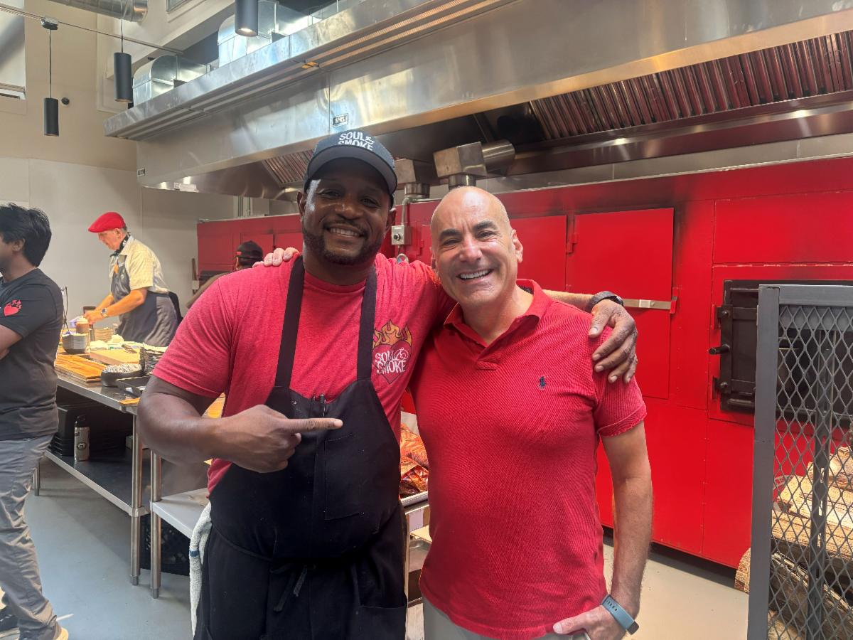 Two smiling men in red shirts stand with their arms around each other in a restaurant kitchen. One wears a black apron and Soul & Smoke cap and points toward the other man; red smokers and kitchen staff are behind them.