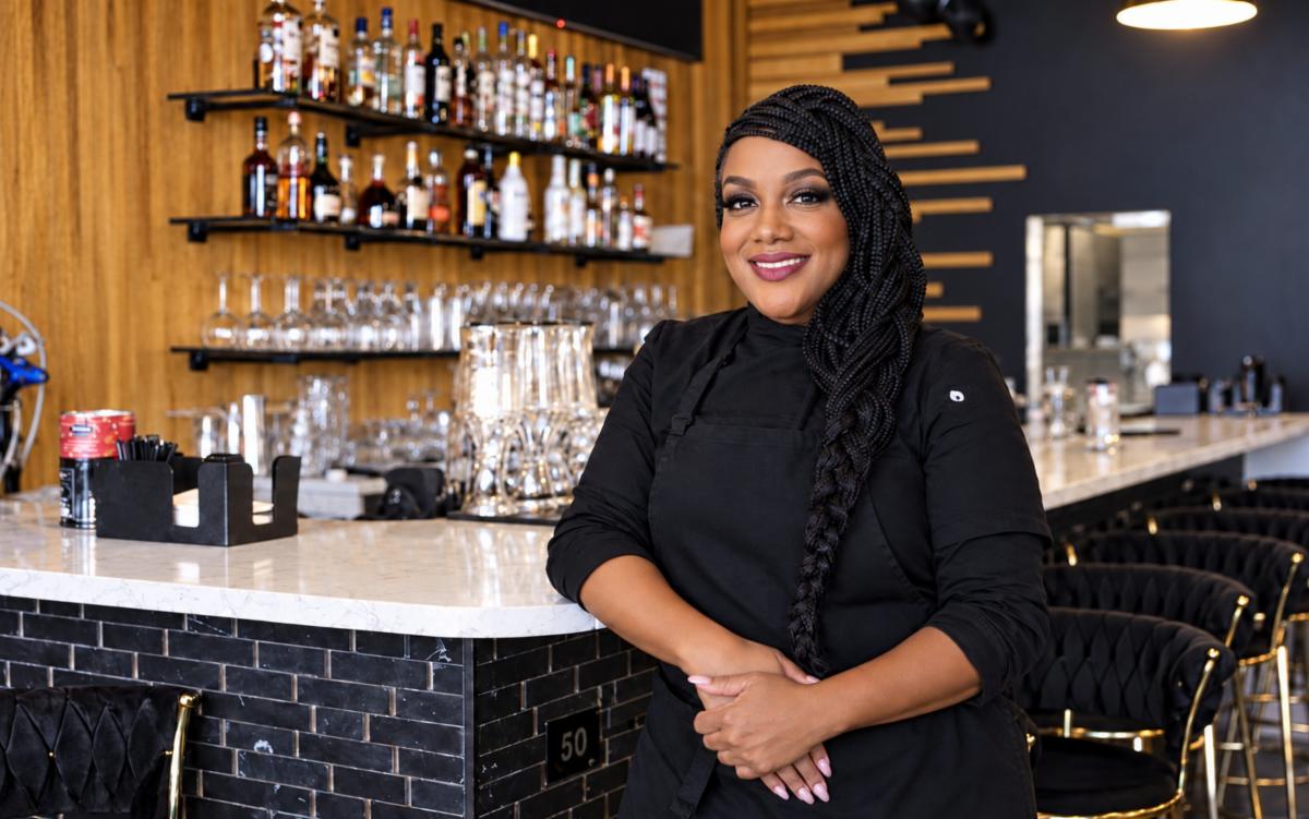 Smiling woman with long black braids wearing a black chef's uniform stands at a marble bar counter with shelves of liquor bottles and glassware behind her.