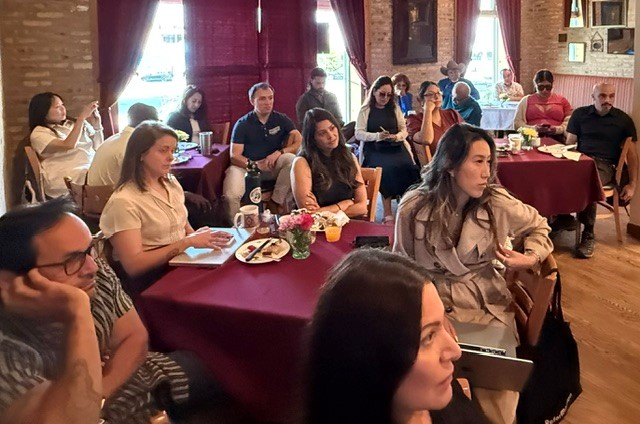 Attendees seated around red-clothed tables in a warmly lit restaurant, listening during a group gathering; plates, drinks, notebooks, and flowers are on the tables.