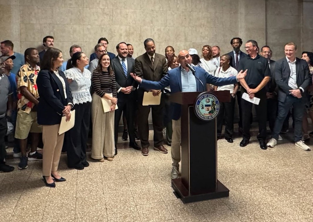 A man speaks at a podium bearing the City of Chicago seal, arms outstretched, as a large group of people stands behind him in a municipal building.