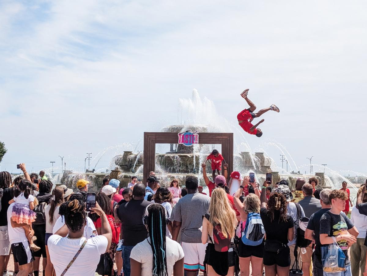 Crowd gathers around Buckingham Fountain at Taste of Chicago as an acrobat in a red outfit performs a high backflip above the audience. Spectators watch and record the performance with the fountain spraying in the background.
