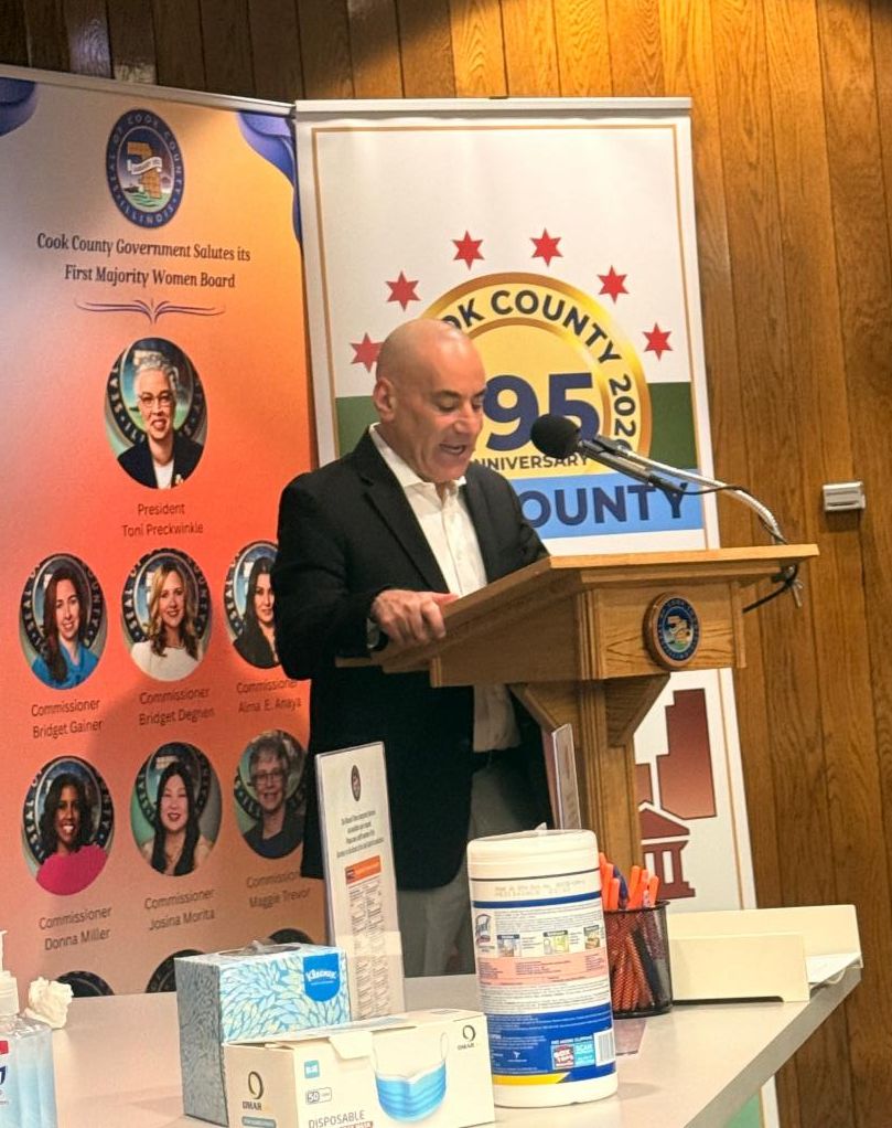 A man in a black suit speaks at a Cook County podium with banners behind him celebrating the county's 195th anniversary and its first majority-women board.