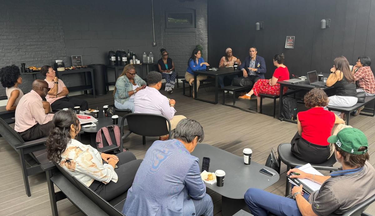 Group of adults seated around tables on a rooftop patio, listening to a man speak during a casual discussion. Coffee cups, laptops, notebooks, and refreshments are arranged throughout the space.