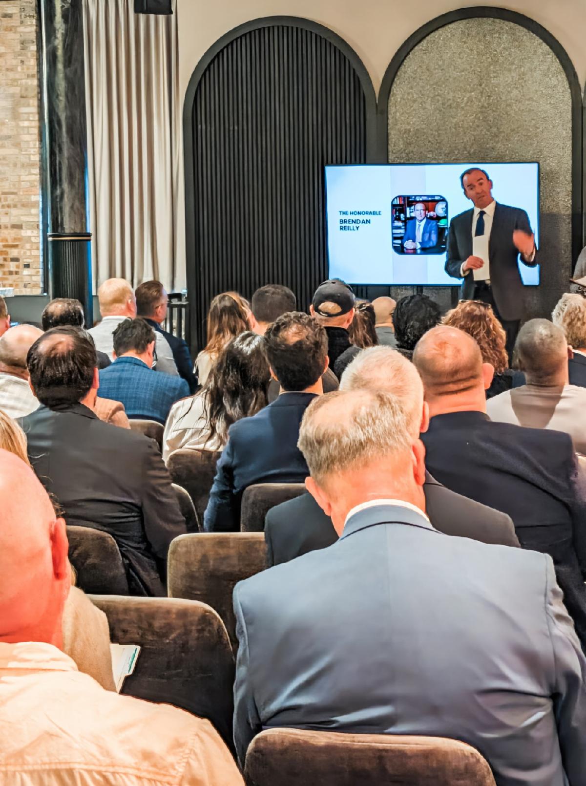 A speaker in a dark suit addresses a seated audience at a professional event, standing beside a screen displaying a presentation featuring The Honorable Brendan Reilly.