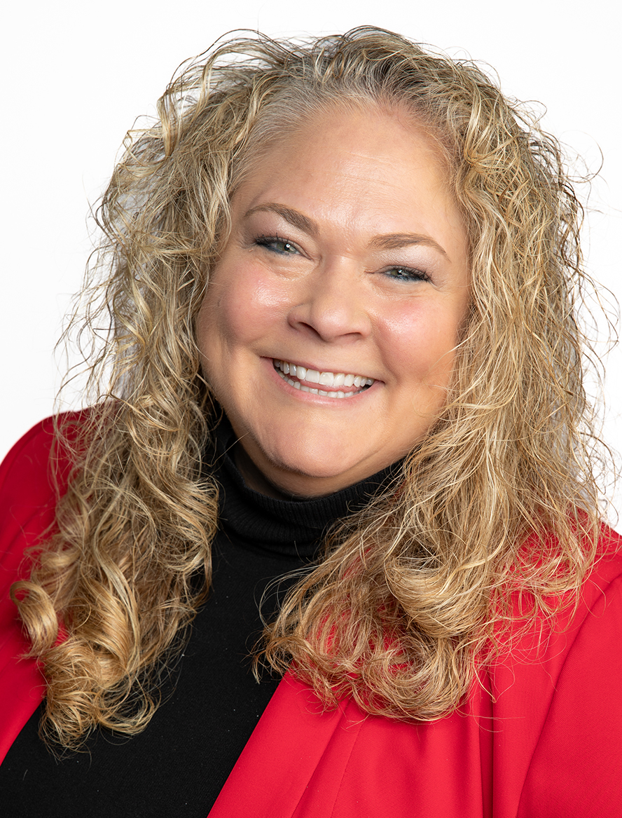 Smiling woman with long curly blonde hair, wearing a bright red blazer over a black turtleneck against a plain white background.
