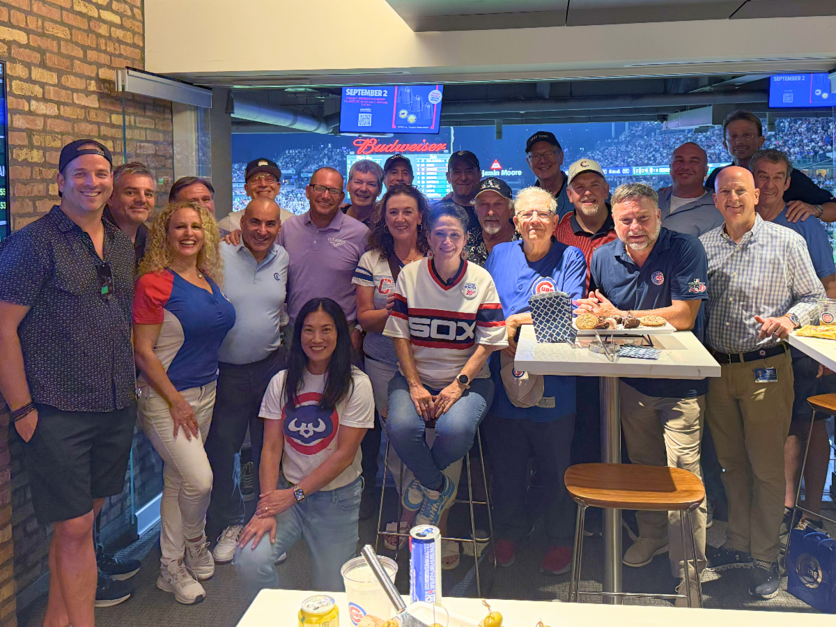 Group of people posing together in a stadium suite overlooking a baseball field with several wearing Chicago Cubs and White Sox apparel.