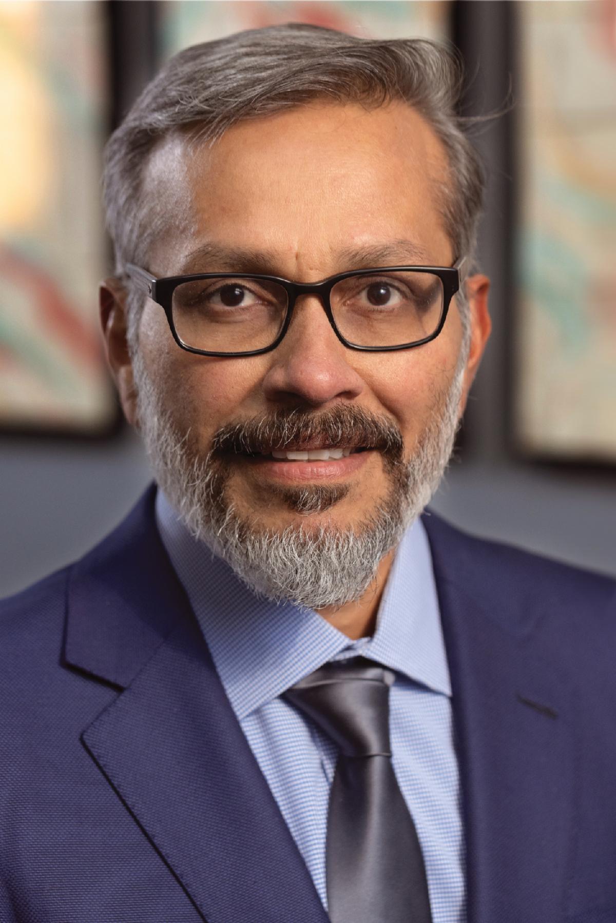 Professional headshot of a smiling man with short gray hair, a salt-and-pepper beard, and black rectangular glasses, wearing a navy suit, light blue shirt, and dark tie against a softly blurred background.