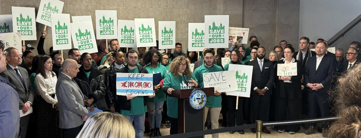 Crowd gathered at a Chicago press conference with speakers at a podium and many people holding signs that read Save Our Jobs, showing restaurant workers and supporters advocating for jobs and local restaurants