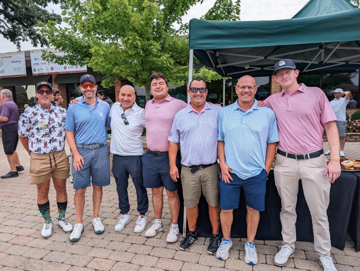 Seven men stand arm in arm outdoors at a golf event, wearing golf shirts, shorts, and caps with trees, sponsor banners, and a green canopy behind them.