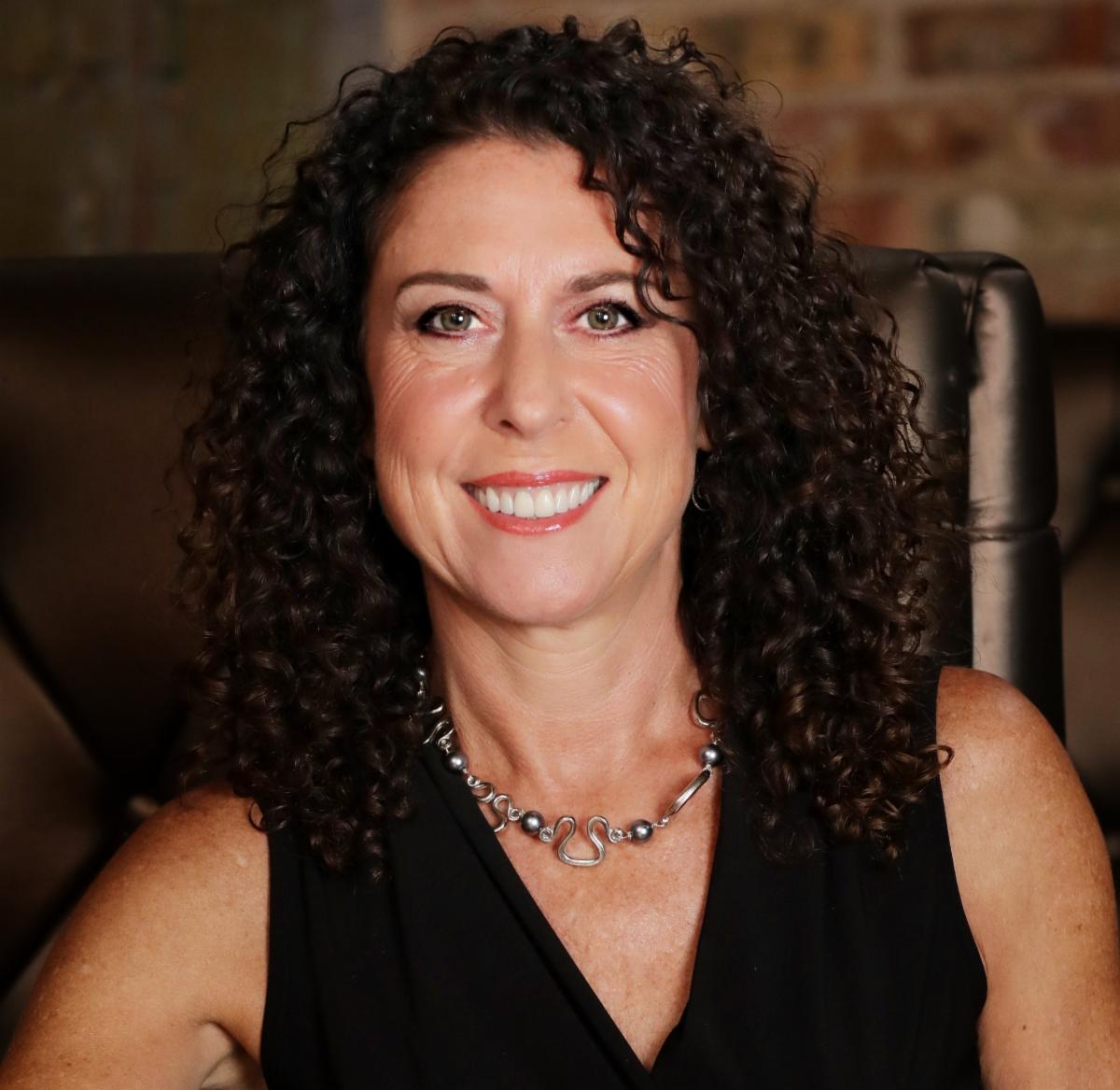 Smiling woman with long dark curly hair wearing a black sleeveless top and silver necklace. She is seated in a black chair against a softly blurred brick background.