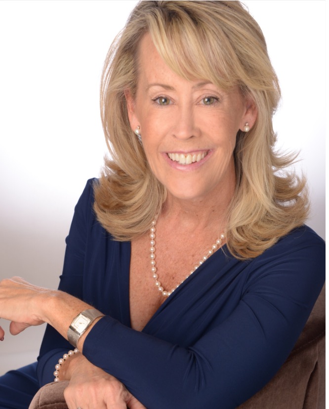 Smiling woman with shoulder-length blonde hair wearing a navy blue dress, pearl necklace, and earrings, seated against a light background with her arms resting on a chair.