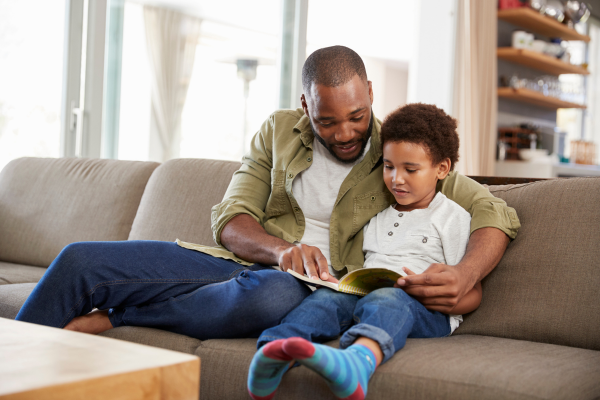 A man and a young child sit together on a couch in a cozy living room. The man is reading a book, holding it open on his lap, while the child attentively looks at the pages. Shelves with various items are visible in the background.