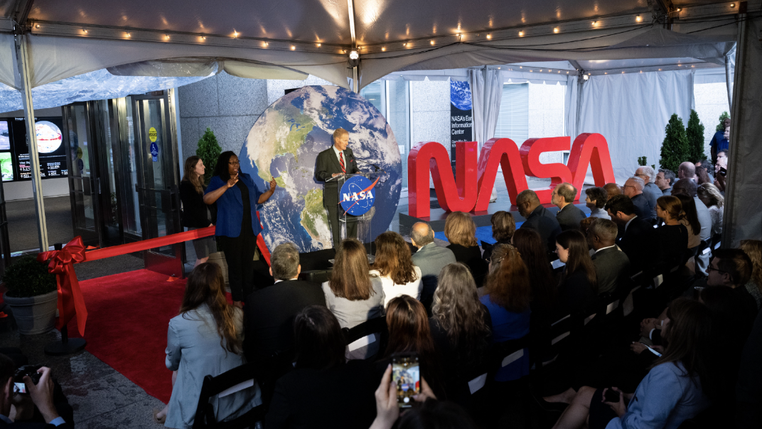 Beneath a white tent in front of the NASA lobby doors and the Earth Information Center, NASA Administrator Bill Nelson stands behind a NASA podium, with a round Earth behind and a NASA worm logo beside him. There is a sign language interpreter signing nearby and a seated audience.