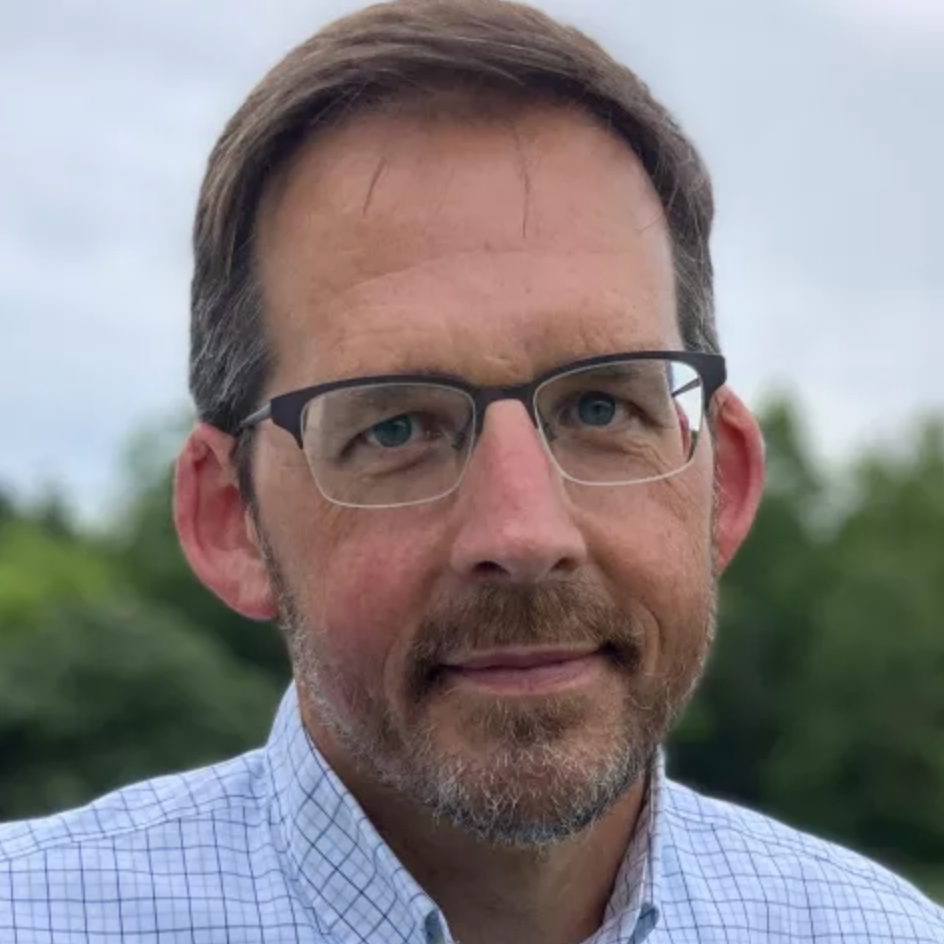 Close-up of Lawrence Friedl’s face looking at the camera with a friendly expression and green trees and a blue sky in the background.