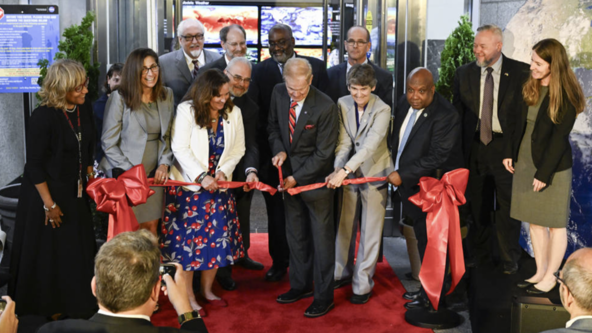 Surrounded by 12 colleagues, Bill Nelson, NASA Administrator, stands in the center of the red carpet, using a large pair of scissors to cut the ribbon at the Earth Information Center's ribbon-cutting ceremony.