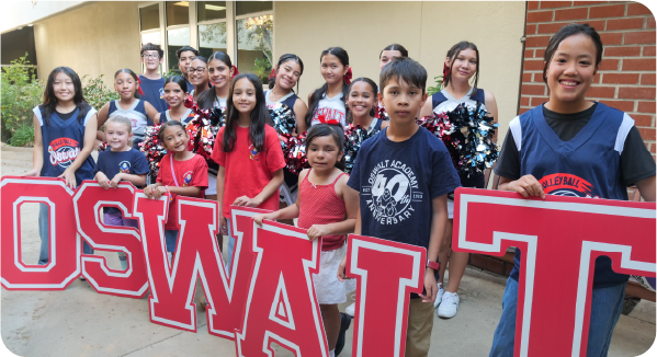 A group of students and young athletes pose outside a school while holding large red-and-white “OSWALT” letters.