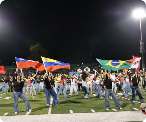 Students perform on a football field at night while carrying colorful international flags, including flags representing Brazil and other countries.