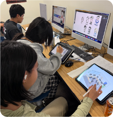 Students sit together at computers and tablets in a classroom, drawing and designing digital illustrations. Two students in the foreground work on tablets while monitors display character drawings and a digital layout.